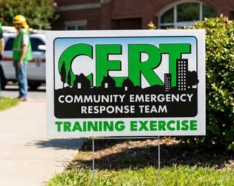 May include: A rectangular yard sign with a white background and green border. The text reads CERT COMMUNITY EMERGENCY RESPONSE TEAM TRAINING EXERCISE in black and green lettering, featuring a silhouette of houses and a building.