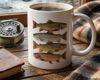 May include: A white ceramic mug featuring a vertical stack of four colorful fish illustrations. Next to the mug sits a brown leather-bound fly fishing journal and a metal fly fishing reel. The mug contains a dark liquid with visible steam.
