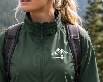 May include: A dark green windbreaker jacket featuring a full-length front zipper, a high collar, and a white graphic print of mountain peaks on the left chest. The jacket is worn with a black backpack with adjustable shoulder straps.