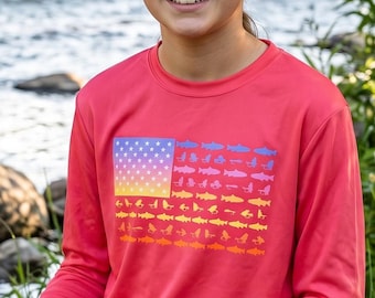 May include: A long-sleeve coral pink shirt featuring a rectangular graphic on the chest. The graphic displays a stylized American flag pattern composed of small, repeating fish silhouettes in shades of blue, yellow, and orange, arranged in horizontal rows.