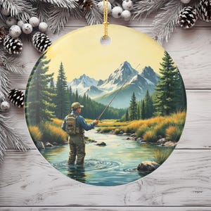 Puede incluir: Adorno navideño redondo de cerámica con una escena de un pescador con mosca en un río. El fondo muestra montañas, árboles y un cielo amarillo. El adorno tiene una cuerda dorada para colgar.