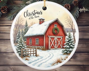 May include: Round Christmas ornament featuring a red barn with snow-covered roof and a green wreath. The ornament has the text "Christmas at the Farm" and a gold string for hanging. The background shows a winter scene with trees and snow.