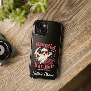 May include: Black phone case with a Halloween-themed design. The case features a cartoon ghost wearing heart-shaped sunglasses, the text "Haunted But Hot - Salem Edition", and the name "Nella's Phone". Roses and a crescent moon are also included.