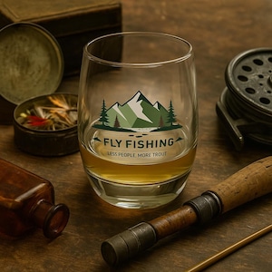 May include: A clear glass with a mountain and tree graphic, and the text "FLY FISHING LESS PEOPLE, MORE TROUT". The glass is filled with a golden liquid. Fishing gear and accessories are in the background.