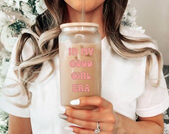 May include: A clear glass tumbler with a wooden lid and a straw, filled with a light brown beverage. The words "IN MY GOOD GIRL ERA" are printed in pink, retro-style lettering on the front of the glass. The tumbler is held by a person.