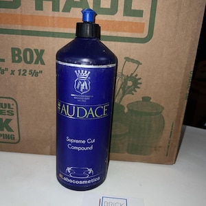 Puede incluir: Una botella azul de pulimento para coches #Audace Supreme Cut Compound con una etiqueta blanca y un logotipo de corona. La botella tiene una tapa azul y está sobre una superficie blanca.