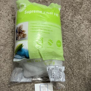 May include: A clear plastic bag filled with white fiberfill stuffing. The bag is labeled "Supreme Fiber Fill" and features a green and white logo with the text "Crafty Threads". The bag also includes a warning label and a net weight of 1/2 (227g).