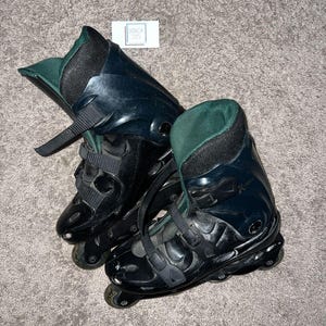 Op de afbeelding: Paar zwarte inline skates met groen en zwart gevoerde binnenkant. De skates hebben verstelbare bandjes en wielen. De foto is van bovenaf genomen.