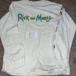 May include: White long-sleeve shirt featuring the text "Rick and Morty" in blue and green. The shirt has character graphics on the right sleeve. The shirt also has the text "Rick and Morty" printed on the collar.