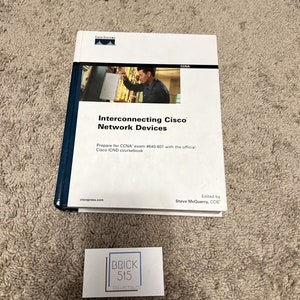 May include: A white book with a blue spine titled "Interconnecting Cisco Network Devices". The book cover features a photo of a person working on a network device. The book is published by Cisco Press and edited by Steve McQuerry, CCIE. A business card for Brick 515 Collectibles is also visible in the image.