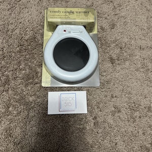May include: White electric candle warmer with an on/off switch. The warmer is packaged in a clear plastic box with the text "comfy candle warmer" printed on the front. Warms candles to release their scent without burning.