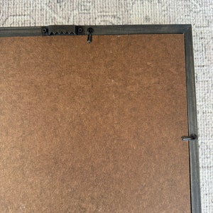 May include: A brown wood picture frame with a black painted border. The frame has a metal hanging hook on the back.