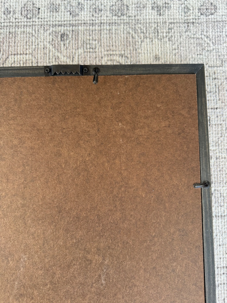 May include: A brown wood picture frame with a black painted border. The frame has a metal hanging hook on the back.