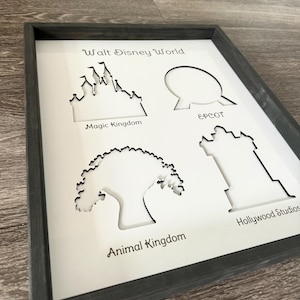 May include: A white framed print with black cutouts of the four theme parks at Walt Disney World. The cutouts are of Cinderella's Castle, Spaceship Earth, the Tree of Life, and the Hollywood Hills. The text "Walt Disney World" is at the top of the print, and the names of each park are below their respective cutouts.