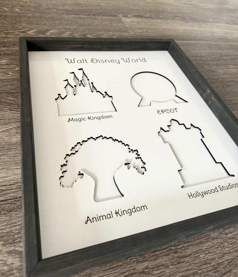 May include: A white framed print with black cutouts of the four theme parks at Walt Disney World. The cutouts are of Cinderella's Castle, Spaceship Earth, the Tree of Life, and the Hollywood Hills. The text "Walt Disney World" is at the top of the print, and the names of each park are below their respective cutouts.