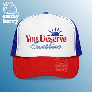 May include: A white foam front trucker hat with a red brim and blue mesh back. The front features the text You Deserve Sunshine in red and blue lettering, accompanied by a blue graphic of a rising sun icon above the word Sunshine.
