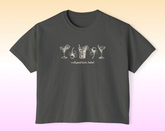 Op de afbeelding: Donkergrijs t-shirt met een witte afbeelding van vier cocktailglazen en klavers. De tekst "well good luck, babe!" staat eronder. Het shirt heeft korte mouwen en een ronde hals.