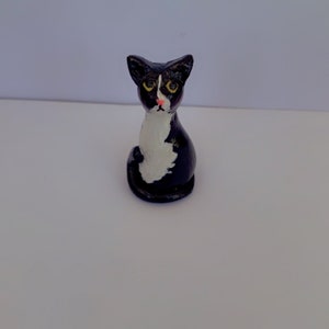 Könnte beinhalten: Kleine handgefertigte Katzenfigur aus Keramik. Die Katze ist hauptsächlich schwarz mit weißen Markierungen auf Brust und Gesicht. Sie hat gelbe Augen und eine rosa Nase. Die Katze sitzt aufrecht, mit dem Schwanz um den Körper.