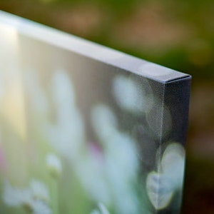 May include: Close-up of a canvas print corner. The canvas is stretched over a wooden frame, with a dark edge. The image on the canvas is a blurred, out-of-focus nature scene with green and white tones.