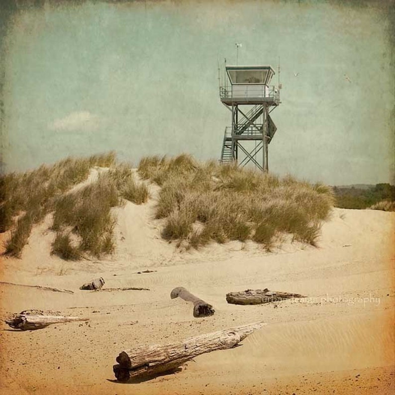 May include: A vintage-style photograph of a tall, metal observation tower on a sandy beach with tall grass dunes. The tower has a white roof and a ladder leading up to the top. There are several pieces of driftwood scattered on the sand.