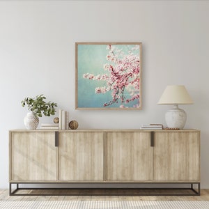May include: A framed print of a pink cherry blossom branch against a blue and green background. The print is hanging on a white wall above a light wood cabinet with two doors.