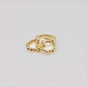 Peut inclure: Trois bagues dorées à perles empilées les unes sur les autres. La bague du haut a une perle blanche avec la lettre "L" gravée dessus.