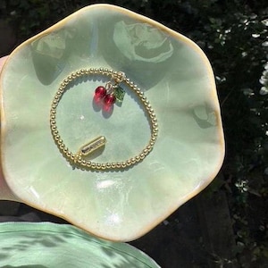 May include: A gold beaded bracelet with a charm of two red cherries and a green leaf. The bracelet is on a green ceramic dish.