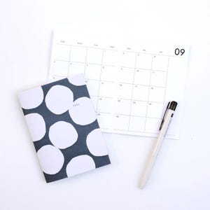 May include: A dark gray notebook with a white circular pattern and the year 2027 printed on the cover, placed next to an open monthly calendar page for September with the number 09 in the corner and a white retractable ballpoint pen.