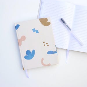 Puede incluir: Un cuaderno color crema con un patrón abstracto colorido de formas azules, rosas y marrones. El cuaderno está cerrado con un marcapáginas de cinta morada clara. Un cuaderno abierto con páginas rayadas y un bolígrafo están al lado.