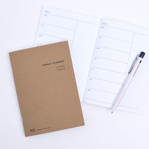Puede incluir: Una agenda semanal con cubierta de papel marrón con el texto WEEKLY PLANNER 52 WEEKS UNDATED y DOZI made in the usa impreso en la parte frontal. Incluye una agenda abierta con páginas blancas que muestran los días de la semana y un bolígrafo.