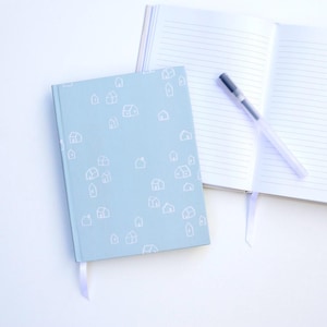 Könnte beinhalten: Ein hellblaues Notizbuch mit einem weißen Hausmuster und einem passenden Lesezeichen aus Band. Ein offenes Notizbuch mit linierten Seiten und einem Stift liegen daneben. Die Notizbücher liegen auf einer weißen Oberfläche.