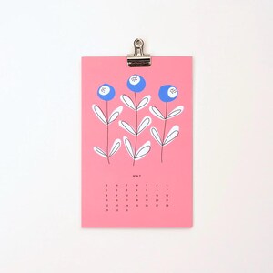 May include: A pink calendar with a blue and white floral design. The month of May is displayed with the days of the week labeled. The calendar is hanging from a silver clip.