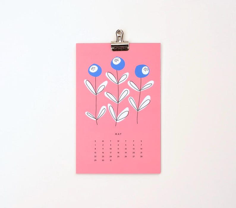 May include: A pink calendar with a blue and white floral design. The month of May is displayed with the days of the week labeled. The calendar is hanging from a silver clip.