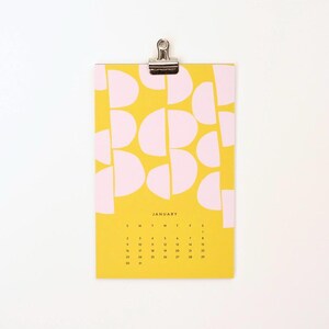 May include: A yellow calendar with a pink geometric pattern. The calendar is for January and has the days of the week labeled at the top. The calendar is hanging from a silver clip.