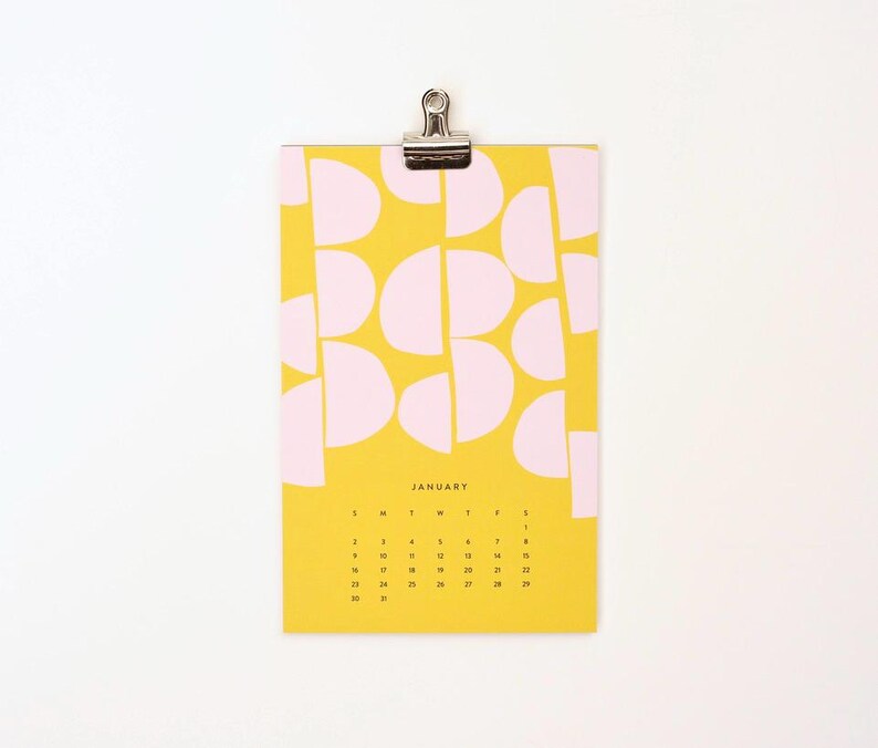 May include: A yellow calendar with a pink geometric pattern. The calendar is for January and has the days of the week labeled at the top. The calendar is hanging from a silver clip.
