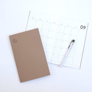 May include: A tan 2027 monthly planner with a closed cover and an open interior page showing a grid calendar for the month of September, labelled with the number 09. A white ballpoint pen rests diagonally across the open calendar page.