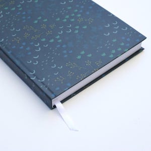 May include: A hardcover journal with a dark blue cover featuring a pattern of small, stylized floral and geometric shapes in shades of blue, green, and gold. A white ribbon bookmark is visible. The journal is on a white surface.