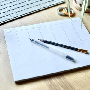 May include: A white weekly planner with a grid pattern, two pens, and a pencil on a wooden surface.