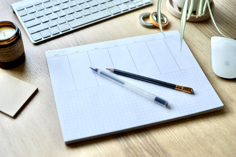 May include: A white weekly planner with a grid pattern, two pens, and a pencil on a wooden surface.