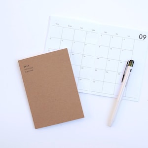 May include: A brown paper-covered 2027 monthly planner notebook resting on an open calendar page with a grid layout for the month of September, accompanied by a white retractable pen with a black clip and a green accent on the top.