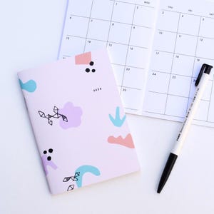 以下が含まれることがあります： 青、ピンク、紫の抽象的な形が描かれた、明るいピンク色の2026年版プランナー。開いたカレンダーと白黒のペンが隣にあります。プランナーは、ミニマリストデザインのソフトカバーです。