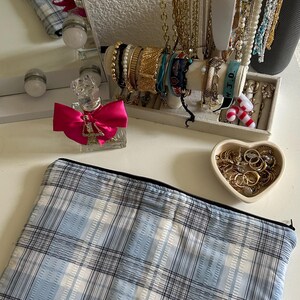 May include: A collection of jewelry and accessories displayed on a white surface. Includes gold necklaces, bracelets, rings, and a clear perfume bottle with a pink bow. A blue and white plaid pouch is in the foreground.