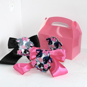 May include: A pink gift box and two satin bows, one black and one pink, each adorned with a cartoon cow. The cows have pink bows and floral accents. The gift box and bows are on a white surface.