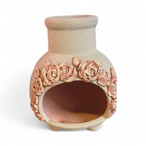 May include: A beige ceramic chiminea with a rounded body, a wide opening, and a short chimney. The chiminea is decorated with raised, pink rose appliques around the opening. It has three small feet for stability.