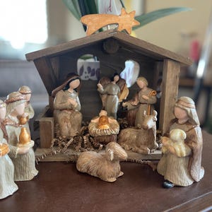 May include: A detailed nativity scene with figurines of Mary, Joseph, baby Jesus, angels, and the three wise men. The figures are in muted tones of beige, brown, and gold, set inside a wooden stable. A wooden star sits atop the stable.