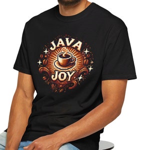 Op de afbeelding: Zwart t-shirt met een koffiekopje-afbeelding en de woorden "JAVA JOY" in een cirkelvormig ontwerp. De afbeelding is in bruin- en goudtinten. De persoon draagt een lichtblauwe spijkerbroek en zit op een zwarte kruk.