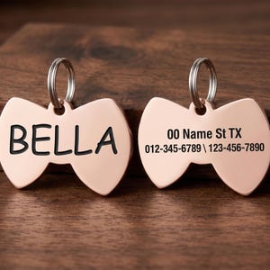 May include: Two rose gold pet tags in a bow tie shape. One tag has the name "BELLA" in black, the other has address and phone number. Each tag has a silver ring for attachment.