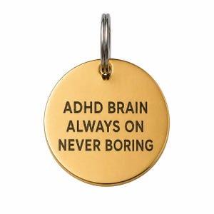 May include: A gold-colored, circular pendant with a silver-colored ring at the top. The pendant has the words "ADHD BRAIN ALWAYS ON NEVER BORING" engraved in black, centered on the surface. The background is white.