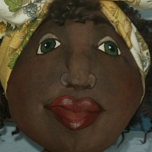 May include: A close-up of a doll's face with brown skin, green eyes, and red lips. The doll is wearing a yellow and white patterned head scarf.