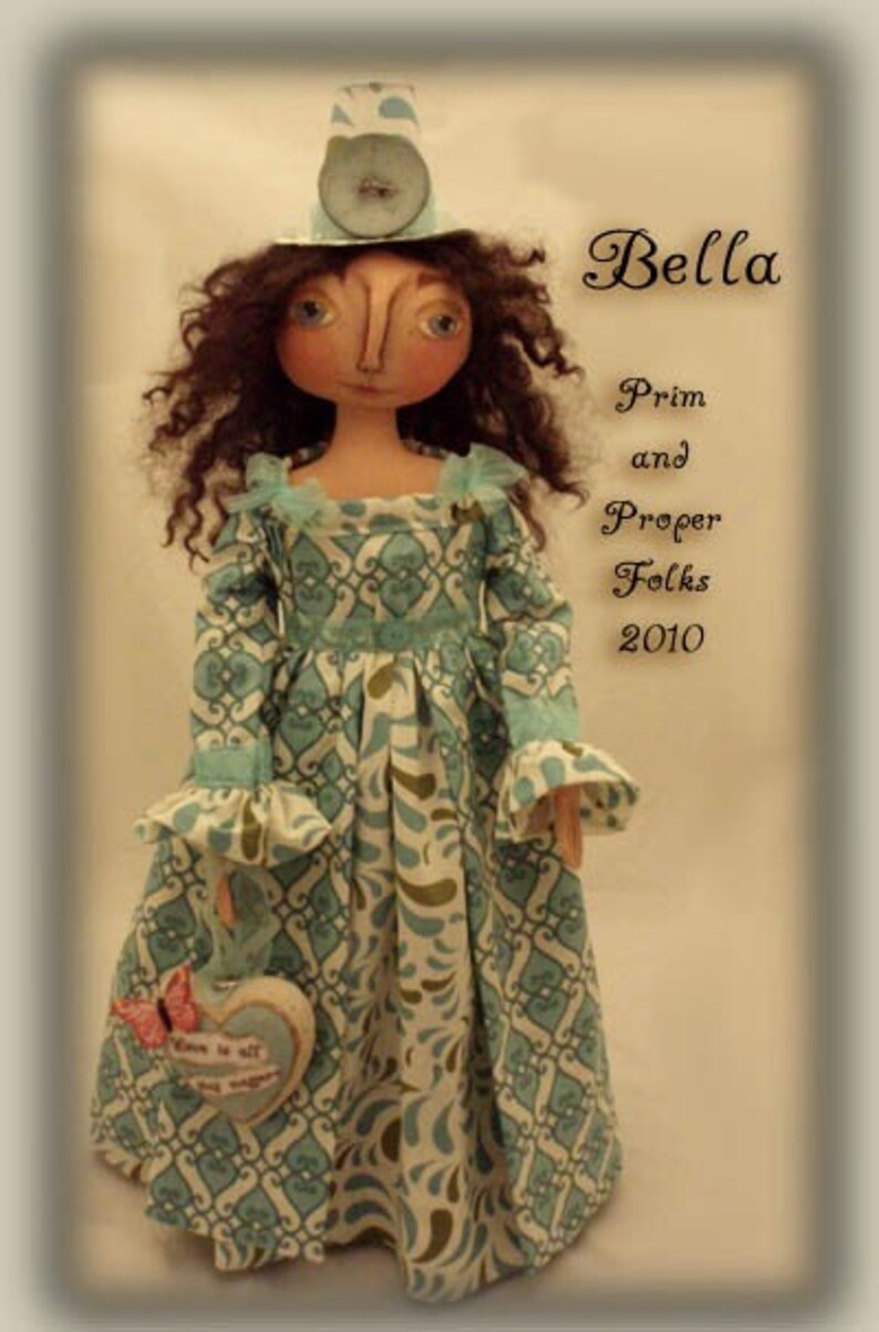May include: A handmade doll called Bella, wearing a blue and white patterned dress and a blue and white hat. The doll is holding a heart-shaped object with the text "Love is all" on it. The doll is part of the Prim and Proper Folks collection from 2010.